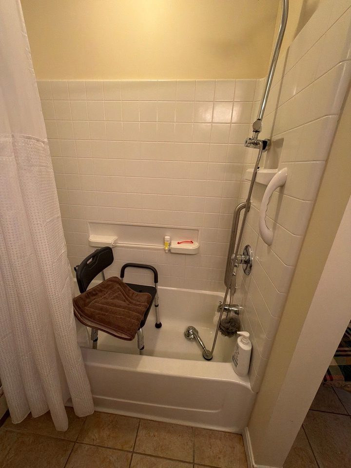 Before: an old white tile tub and shower with a chair and a grab bar